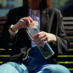 Thirsty young female opening a glass bottle of mineral water to drink. Unrecognizable girl in casual clothes sitting on a bench and drinking fresh water from a reusable glass bottle